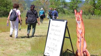 A painted wooden cut-out of a mountain lion sits in the grass next to a whiteboard easel that says "Learn about Water, Wildlife, and Connectivity!" behind which are people walking towards a line of trees
