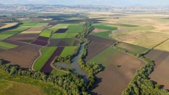 An aerial view of a patchwork of farmlands with a tree-lined creek winding through them; in the far distance are hazy mountains