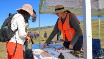 Volunteer wearing a bright orange vest stands behind a table under a pop-up booth and points out something on a map on the table to two visitors who are standing on the other side of the table