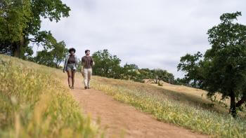 Two hikers wearing brown and tan clothing walk towards the camera on a dirt trail through a yellow grass field with oak trees and hills covered in trees in the background.