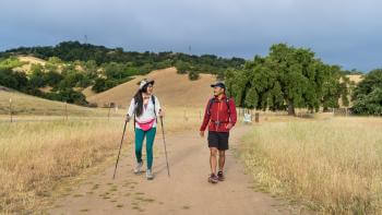 Two hikers walk towards the camera on a dirt trail through a yellow grass field with oak trees and hills covered in trees in the background. The hiker on the left carries hiking poles and wears teal leggings, a pink fanny-pack, and a white jacket. The hiker on the right wears black shorts and a red jacket. They are looking at each other and smiling.