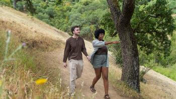 A pair of hikers wearing brown and tan clothes walking down a dirt trail underneath trees towards the camera. The hiker on the right is pointing towards the right of the scene.