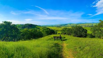 A bench at the top of a green grassy hill overlooking trees and a vast green valley below, with green mountains in the distance. The sky is blue with some light clouds.