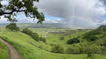 A dirt trail winds through lush green hills dotted with dark green oak trees, in the distance a rainbow arcs over farmland below