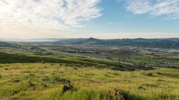 Looking over green grassy hills with orange wildflowers across a valley below towards blue mountains in the distance, under a light blue sky streaked with white clouds