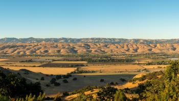 Looking across Coyote Valley, covered in golden fields and farmlands, towards rolling golden hills in the distance all under a blue sky