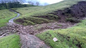 A muddy trail winding through green hills is blocked by a small mudslide cutting across it