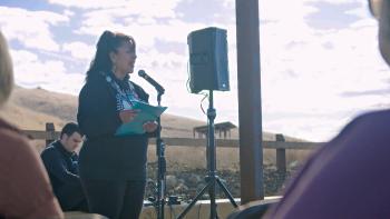 A woman wearing a sweatshirt that says "Muwekma Ohlone Tribe" stands in front of a microphone holding a folder in front of her, delivering a speech. Behind her are golden hills and a cloudy blue sky.