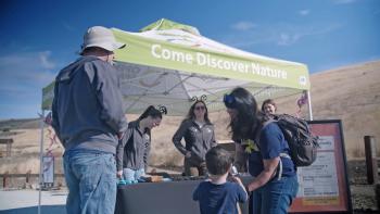 Two employees wearing butterfly headbands stand under a booth tent that says "Come Discover Nature", a family stands in front of the booth