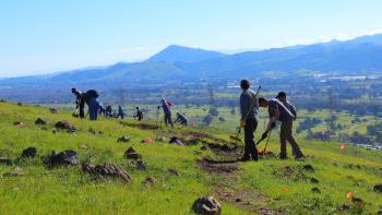 A group of people with shovels and rakes work on a dirt trail winding across a bright green hillside overlooking a valley below and blue mountains in the distance.