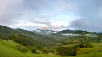 Lush green hills stretching into the distance, covered in green trees, under a light blue morning sky with dark clouds