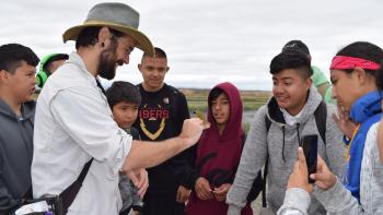 A man with a beard and wide-brimmed hat holds a small bird on his hand, surrounded by teenage students who are smiling and looking at the bird