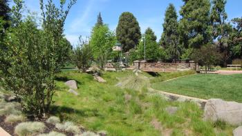 A grassy swale and lawn area with shrubs and small trees, in the distance is a small pedestrian bridge and play structure