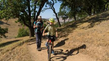 A child bikes on a trail in front of their parent surrounded by trees