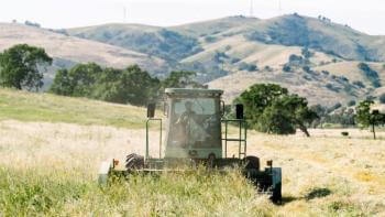 A person on a tractor drives through a hay field with golden hills and trees in the background