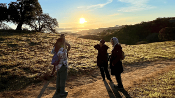 A group of people face each other on a trail during sunrise with hills in the background.