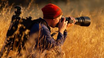 A man wearing a red beanie and navy blue shirt knees in a field of golden grass holding up a camera with a long lens, taking a photo of something to the right