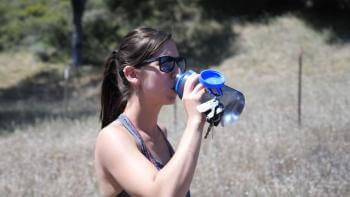 A woman with a ponytail and wearing sunglasses and a gray tank top drinking out of a gray and blue reusable water bottle, in front of a blurry background of brown grass-covered hills with trees