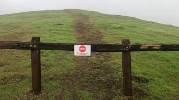 A brown split-rail fence with a small white sign that says "Do Not Enter This Area" in red text below a red stop sign image, in front of a green hill with a faint unsanctioned trail leading up