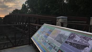 A long educational panel with colorful photos and text boxes in front of a metal and stone pedestrian bridge flanked by trees, under a colorful sunset in the background