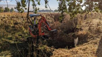 A person on a tractor clears a grassy and overgrown area for installation of a weir