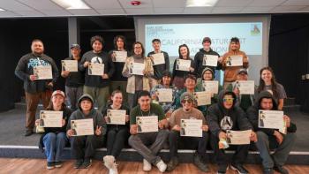 21 young adults sit and stand for a group photo in a classroom setting, holding up certificates and smiling at the camera