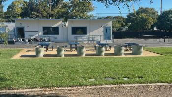 A row of six raised metal garden beds next to four school picnic tables surrounded by green grass, in front of a school blacktop with portable classrooms behind them