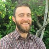 light skinned man with brown hair and full beard against green trees