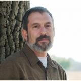 Light skinned man with brown hair and peppered beard wearing a brown button up shirt against a tree