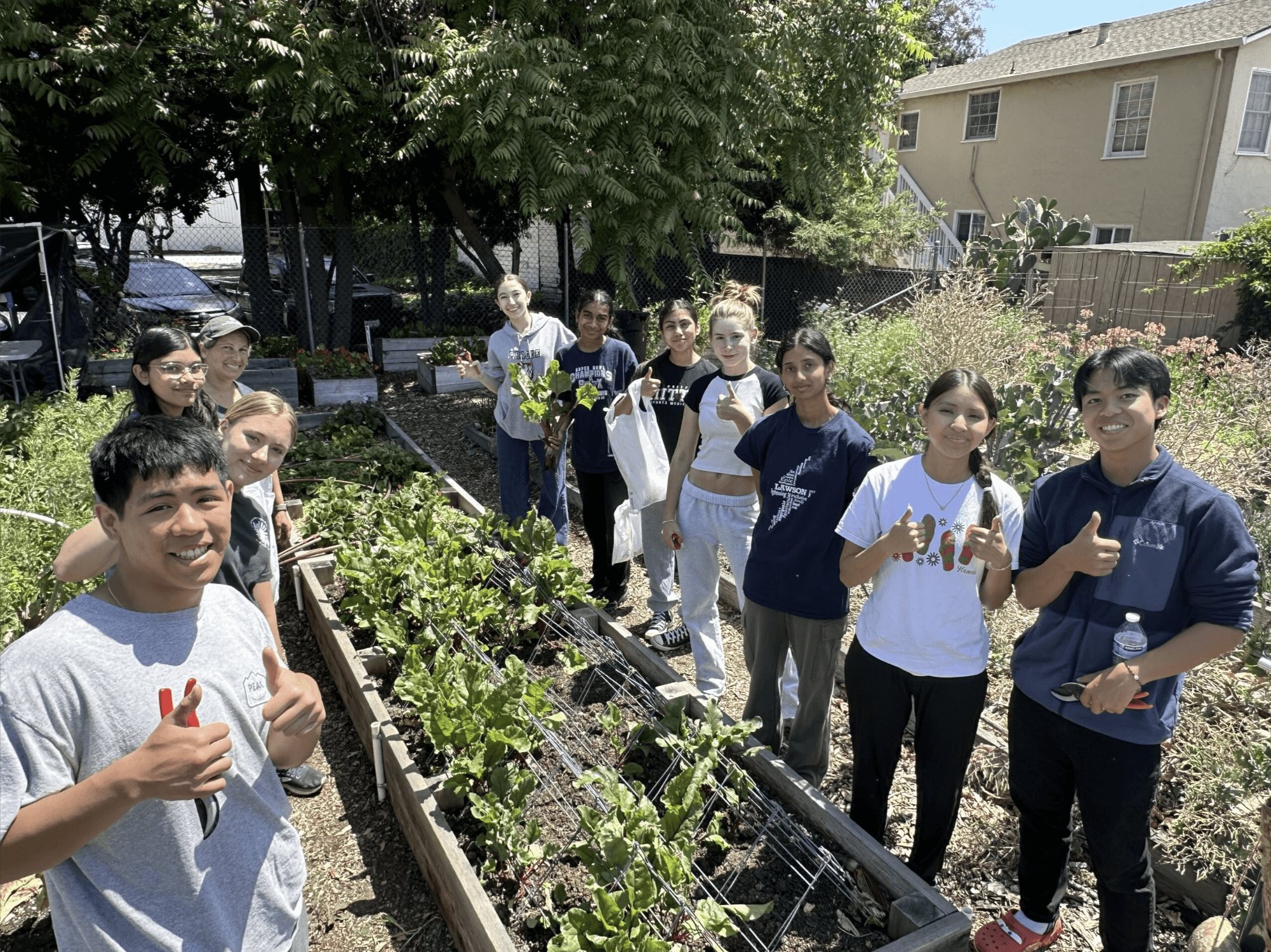 garden box in the center of 2 rows of young people with their thumbs up with trees in the background and a 2 story home on the right side