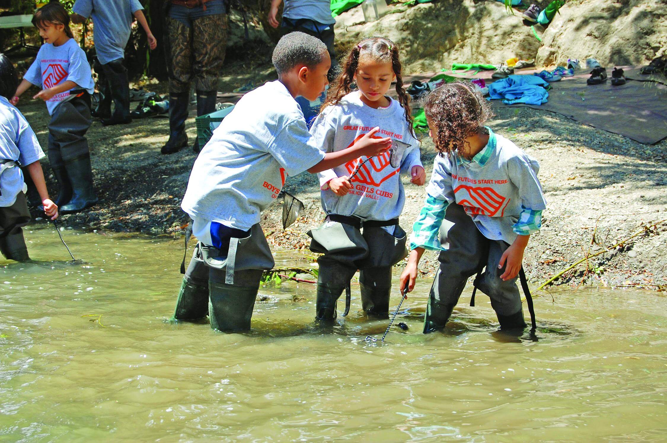 Three children wearing large grey "Boys & Girls Club" t-shirts and waders stand in a full creek holding small nets and looking at something the child on the left is holding up