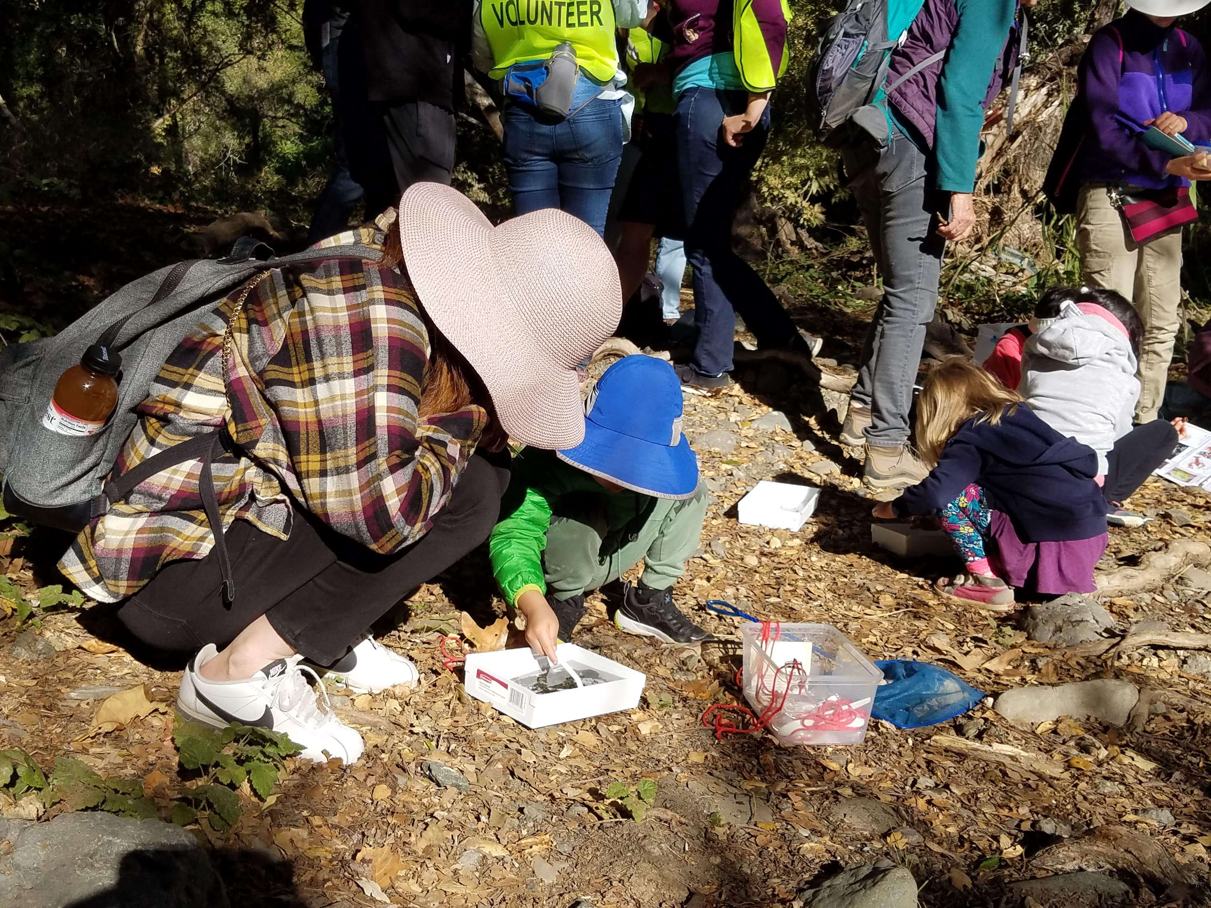 An adult and two children wearing hats squat on the ground and crouch over white square containers filled with water, looking in  