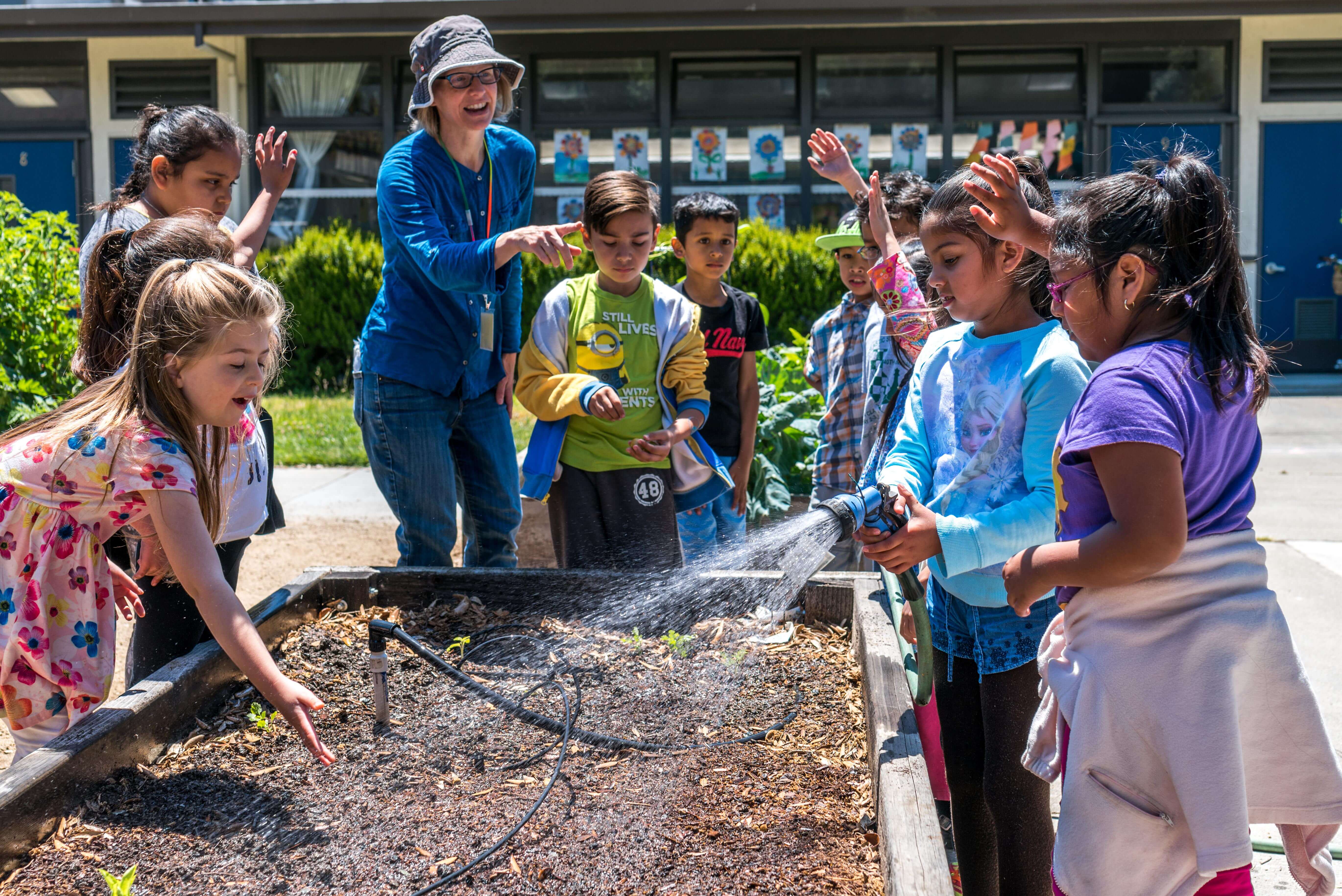 A group of young students in colorful clothing stand smiling and laughing around a raised garden bed, one of the students is holding a sprinkler towards the soil in the bed; a teacher stands to one corner of the bed pointing and smiling at the students