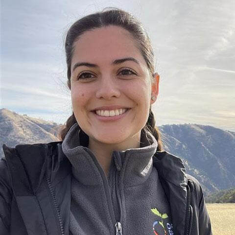 Profile photo of Aline, wearing a gray Open Space Authority jacket and smiling at the camera, with mountains and an open sky in the background.