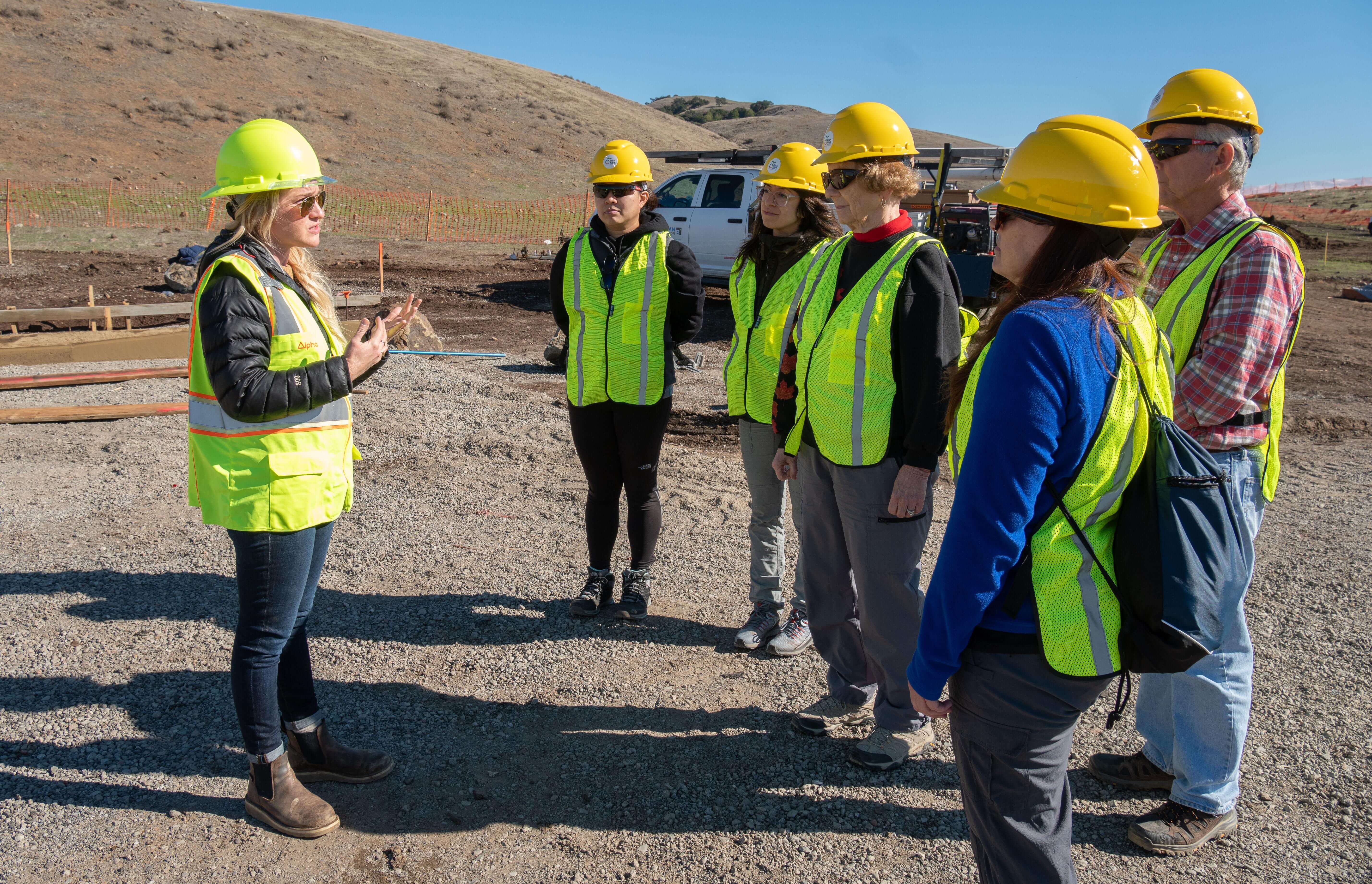 Blonde woman in yellow hard hat and vest speaking to a group also wearing yellow hard hats and vets against a dirt hill an light blue sky