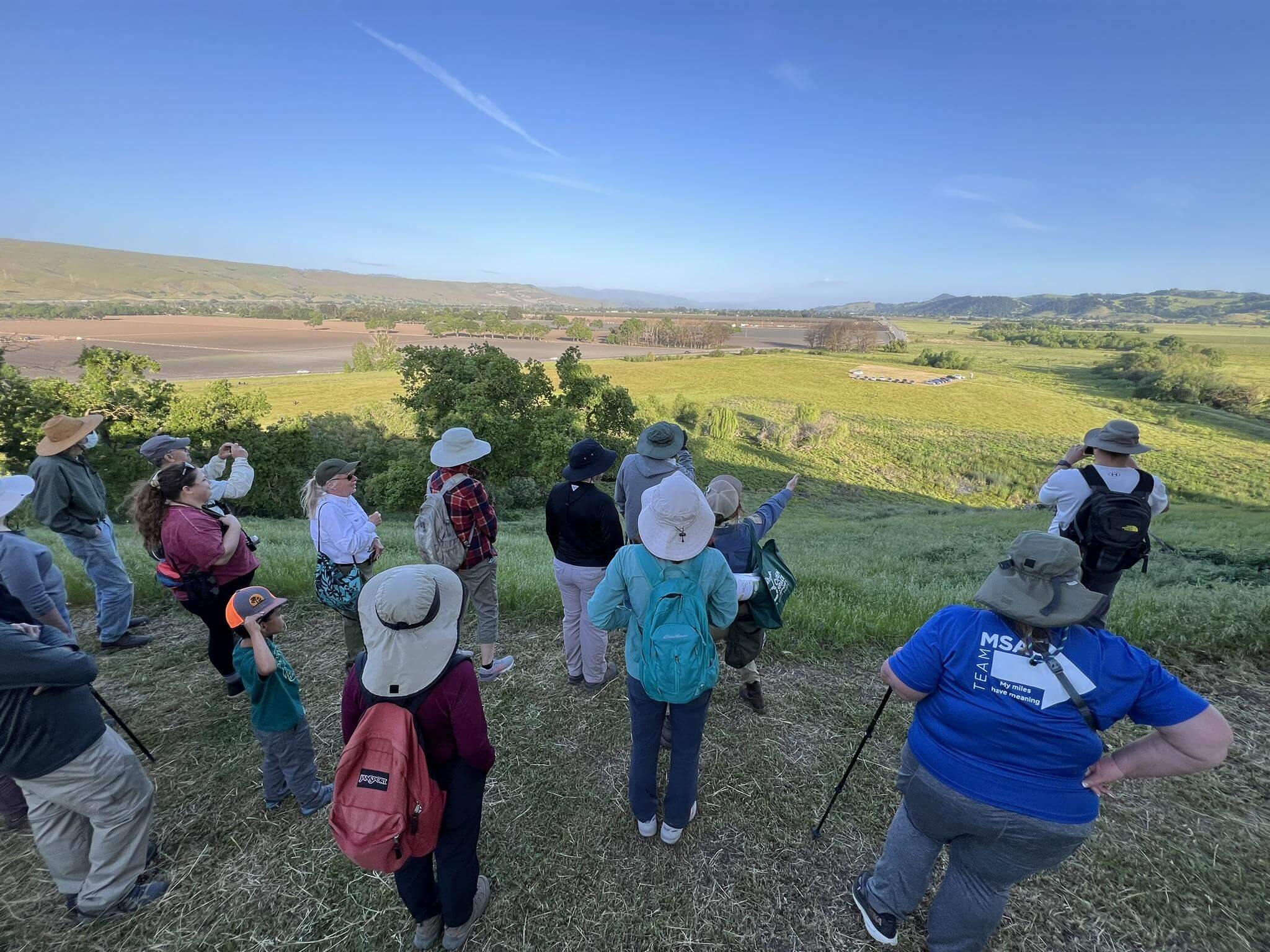 A group of people stand at the top of a hill overlooking a green valley bathed in late-afternoon light, under a clear blue sky