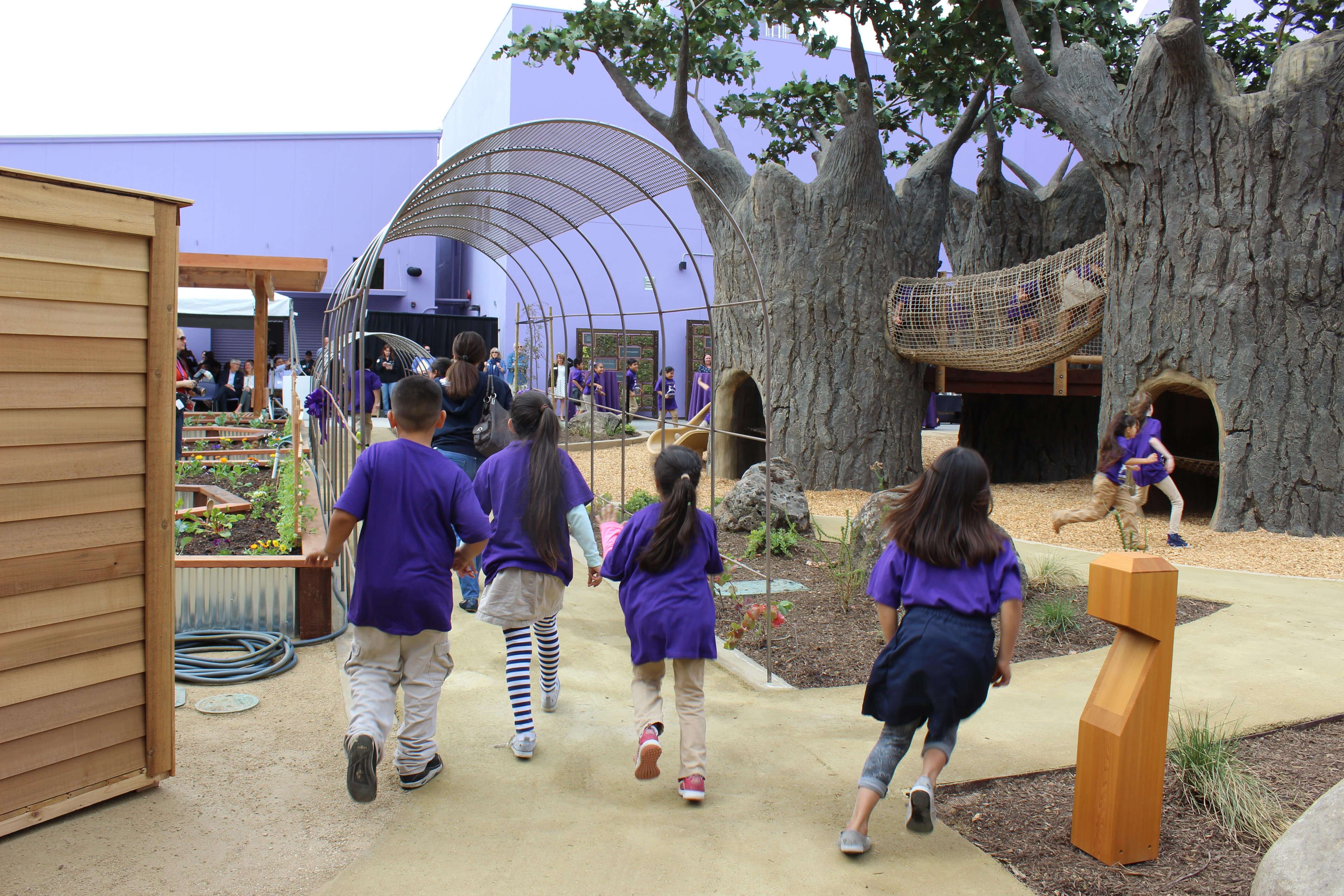 Children wearing dark purple t-shirts running towards an outdoor play area with tree-shaped play structures, a rope bridge, and garden planters