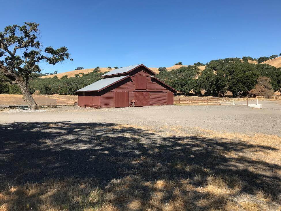 shadow of a large tree in the foreground with a brown barn with a tree to the left and shrub covered hills in the background