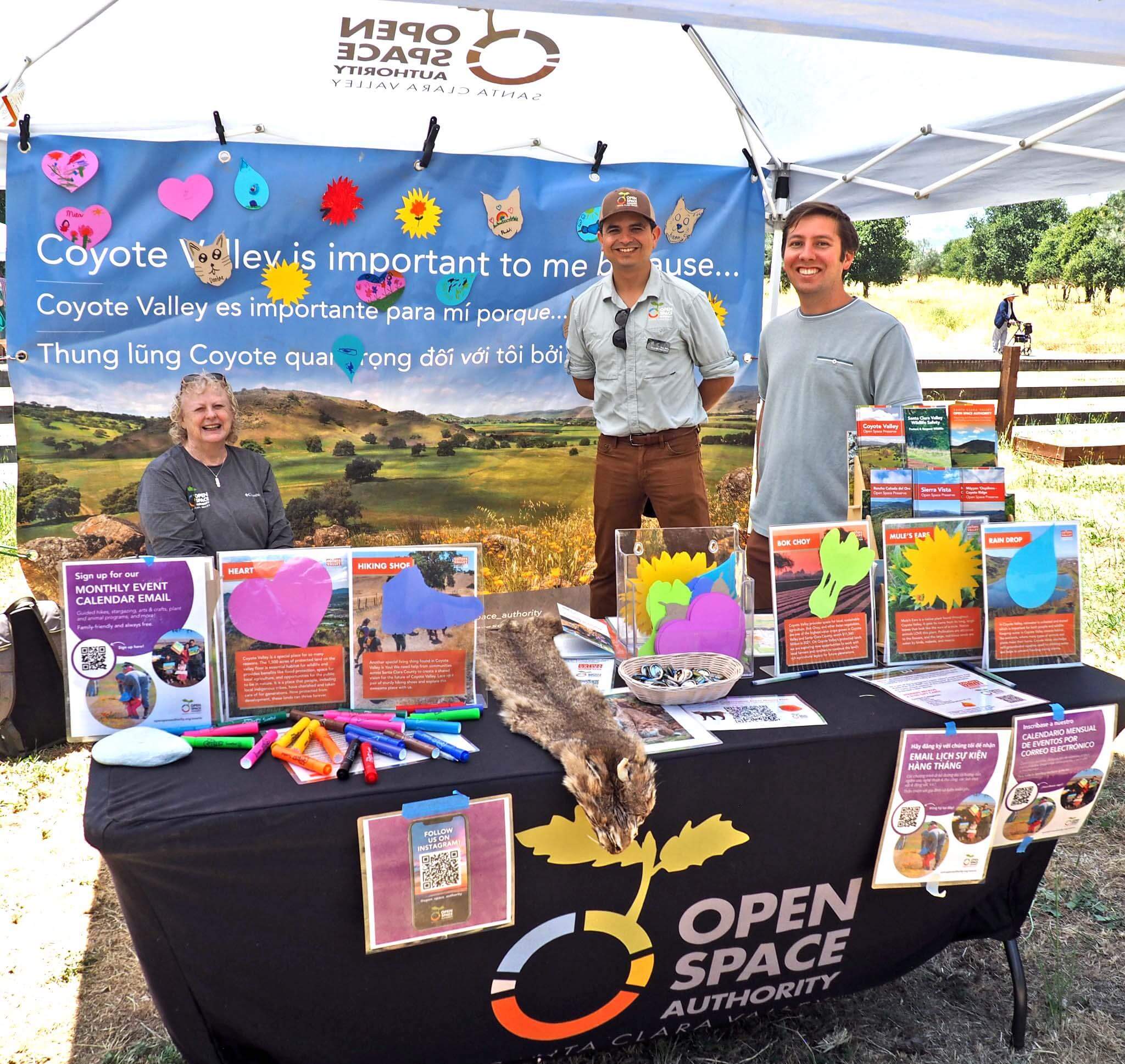 Three people stand smiling at a camera under a booth and behind a table with the Open Space Authority logo and covered in colorful brochures and informational items about Coyote Valley