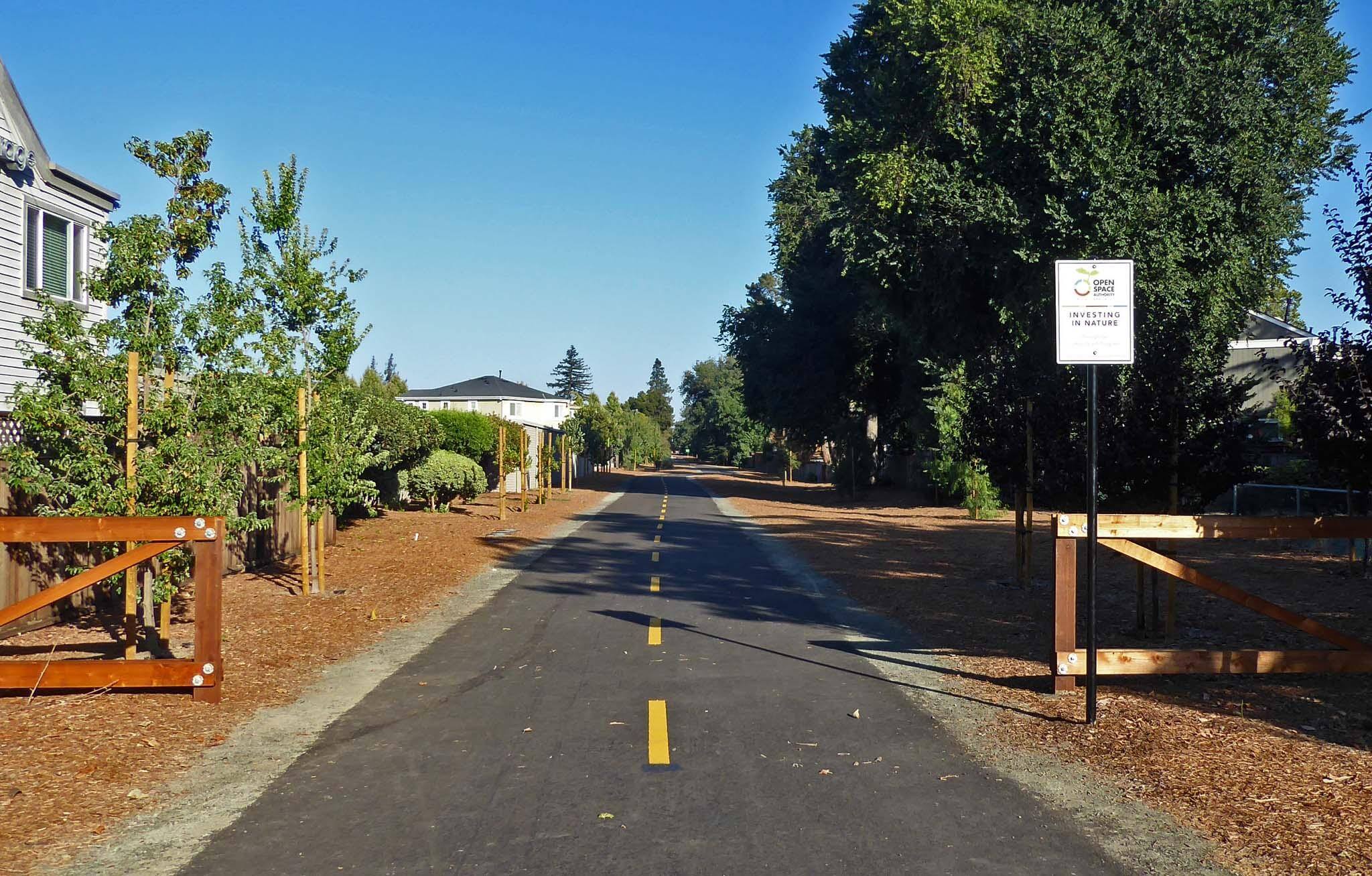 An asphalt walking trail stretching straight away from the camera, bordered by low split-rail fences and bark chip landscaping and trees