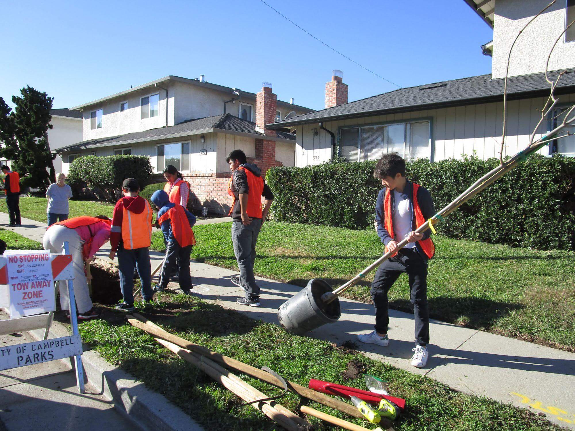 A group of teens wearing orange vests work on planting a tree sapling in a hole next to a sidewalk in a residential neighborhood