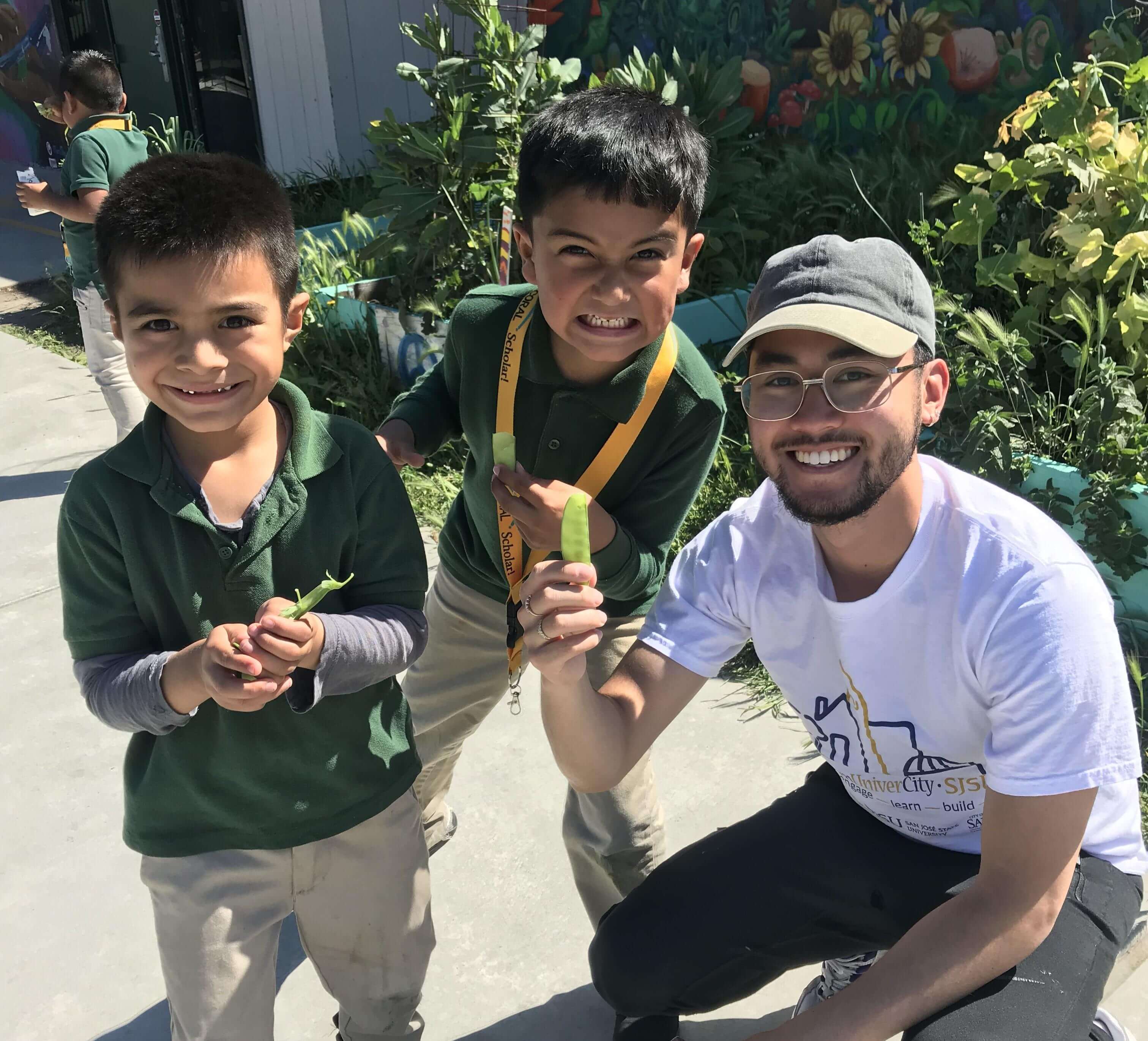 Two young children in green polo shirts stand next to a kneeling young adult in a white t-shirt, holding up pea pods and smiling