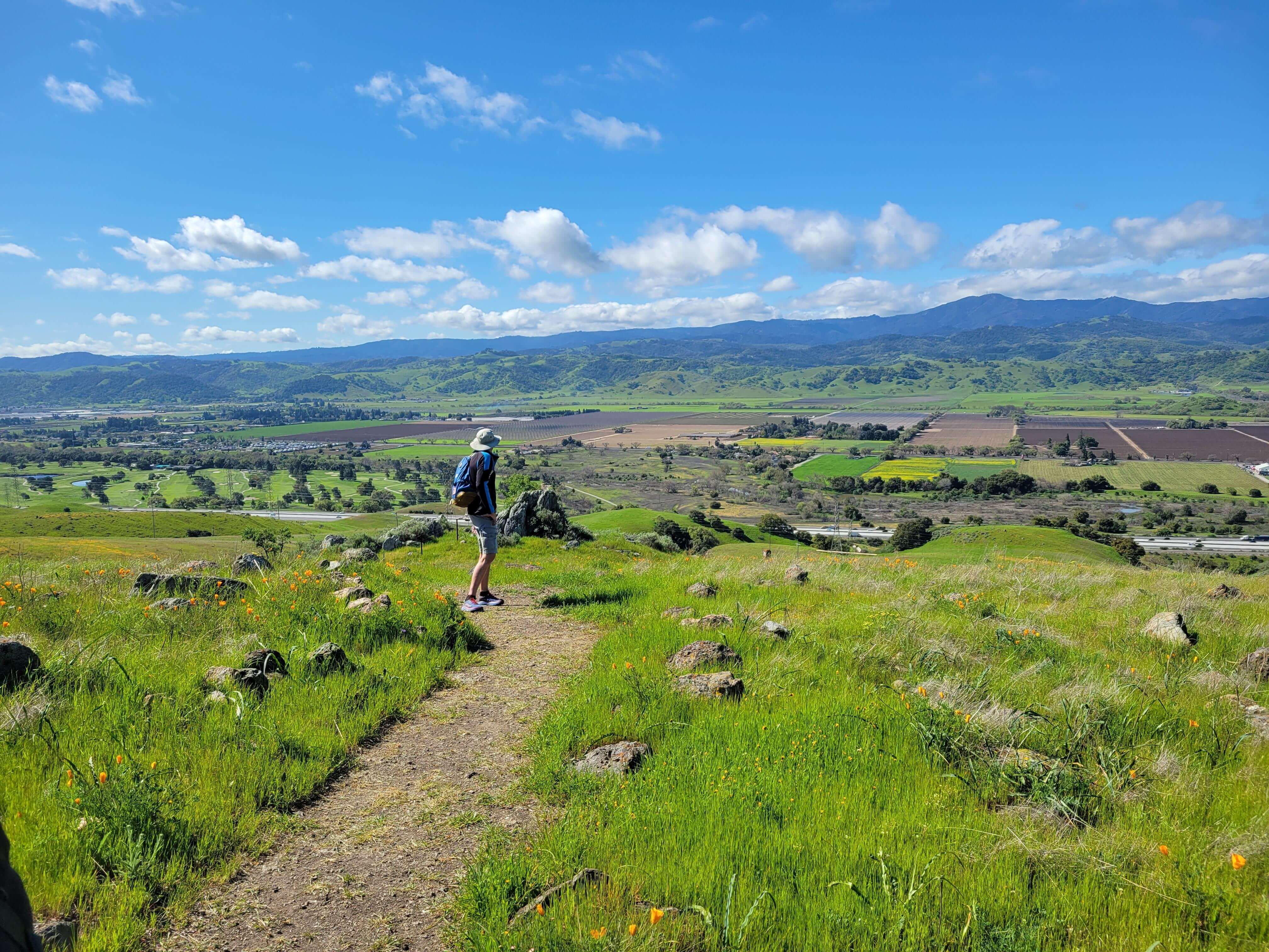 Hiker on a trail on a green grassy field overlooking a beautiful view of a valley patchworked with farm fields and blue mountains in the distance