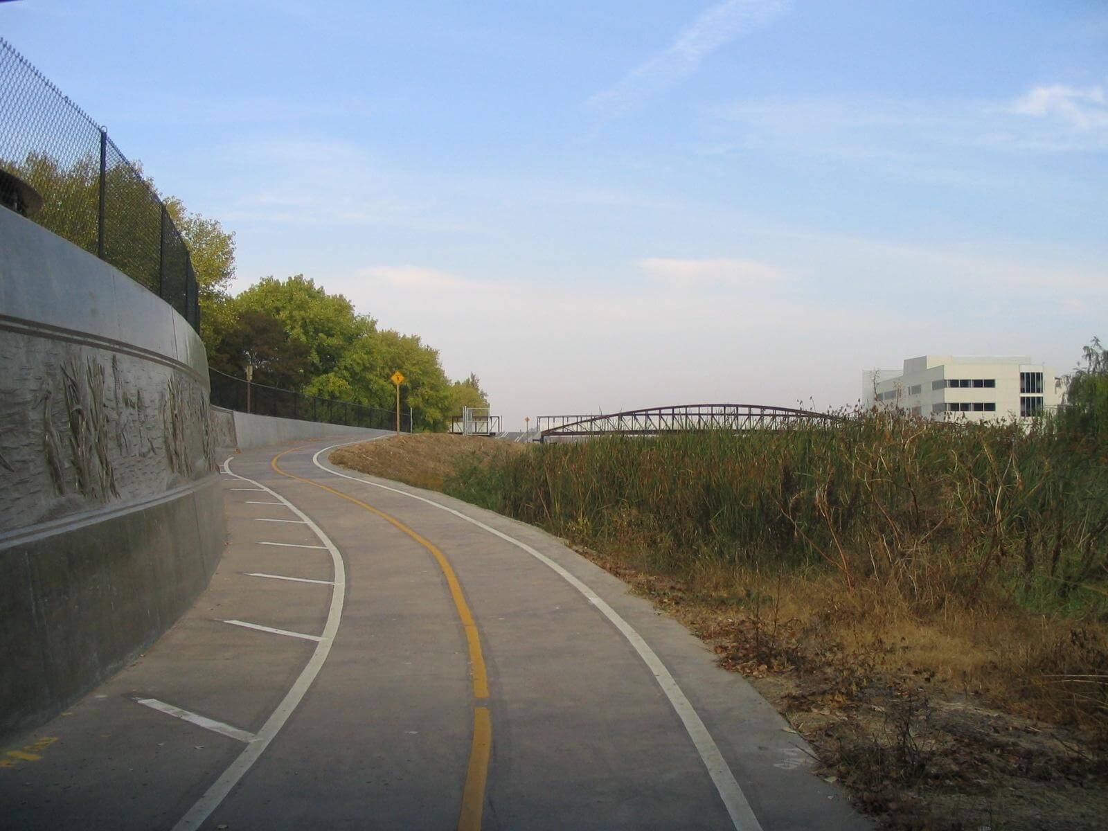 A paved bike trail with yellow and white lines curves up a slight incline with tall grasses on the right and a concrete wall with relief sculptures and a fence top on the left