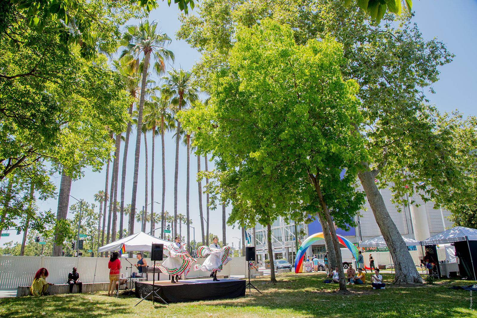 A pair of dancers wearing white traditional Mexican dresses with colorful ruffles perform on a small black stage in a park with tall green trees and festival booths in the background