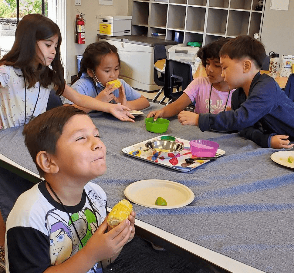 A ground of young students sit around a classroom table, the four in the back reach towards a small colorful dish, one student closer to the camera holds a corn cob in his hands and smiles up to someone our of frame