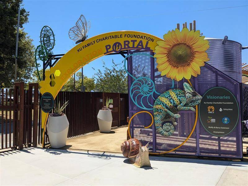 The entrance to an outdoor education area, with a bright yellow arch that reads "Xu Family Charitable Foundation Exploration Portal" surrounded by sculptural and cut-out shapes of a yellow flower, a blue and yellow chameleon, a snail, and a butterfly