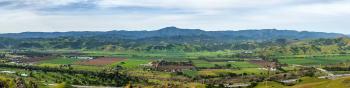 Looking over green hills across a valley below filled with agricultural fields, to blue mountains in the distance, under a blue sky streaked with white clouds 