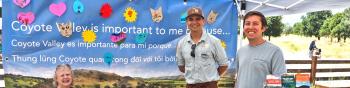Three people stand smiling at a camera under a booth and behind a table with the Open Space Authority logo and covered in colorful brochures and informational items about Coyote Valley