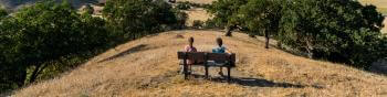 Two hikers sit on a bench overlooking golden hills and trees